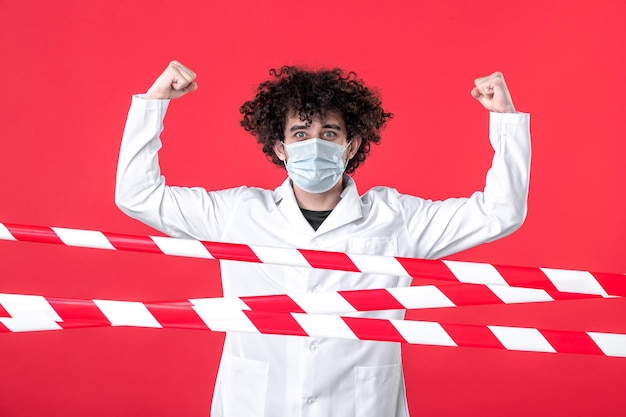 Foto grátis vista frontal jovem médico masculino em uniforme médico e máscara em fundo vermelho perigo covid-quarentena de saúde faixa de aviso hospital