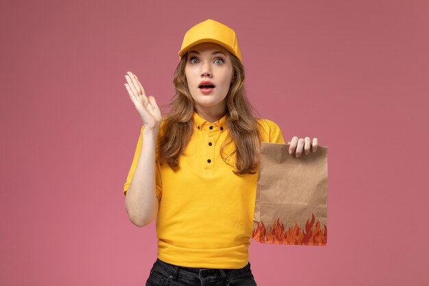 Vista frontal jovem mensageira com capa amarela uniforme amarela segurando pacote de comida no fundo rosa escuro uniforme serviço de entrega de trabalhador feminino