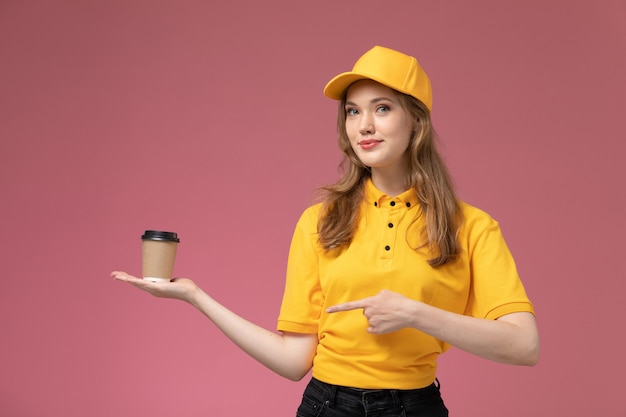 Vista frontal jovem mensageira com capa amarela uniforme amarela segurando uma xícara de café de plástico marrom no fundo rosa escuro.