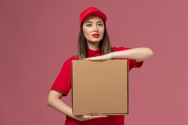 Vista frontal jovem mensageira de uniforme vermelho segurando uma caixa de comida no fundo rosa.