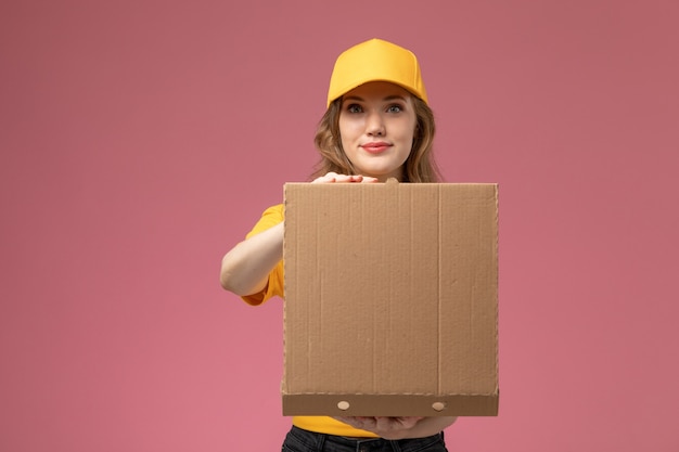 Vista frontal jovem mensageira em uniforme amarelo capa amarela segurando a caixa de entrega de comida abrindo-a no fundo rosa escuro.