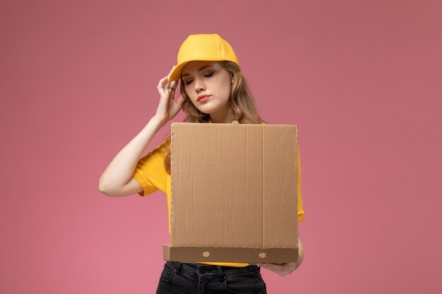 Vista frontal jovem mensageira em uniforme amarelo capa amarela segurando pacote de entrega de comida abrindo-o sobre fundo rosa escuro serviço de entrega uniforme trabalhador feminino