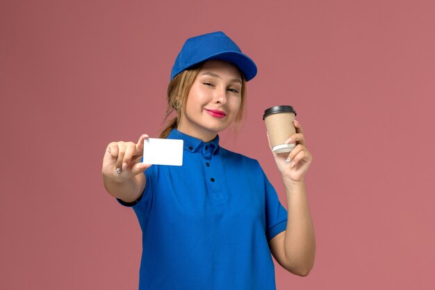 Vista frontal jovem mensageiro de uniforme azul posando segurando xícara de café e um cartão branco com um sorriso, trabalhador de trabalho de mulher de entrega de uniforme de serviço