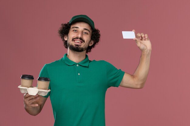 Vista frontal jovem mensageiro masculino em uniforme verde e capa segurando copos de café de entrega e cartão na mesa rosa serviço de trabalho uniforme entregador trabalhador masculino