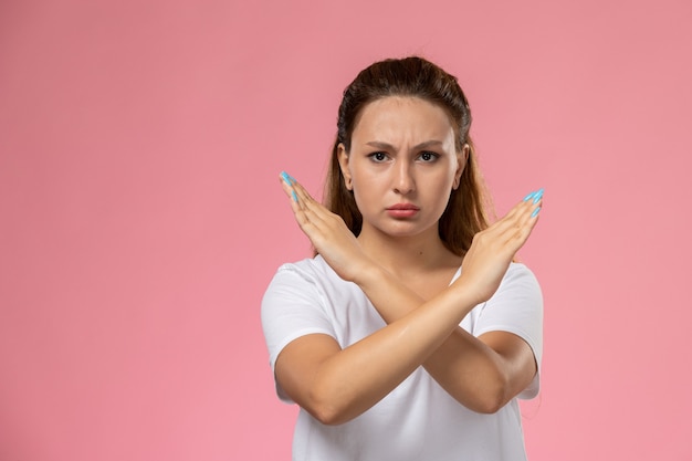 Vista frontal jovem mulher atraente em camiseta branca mostrando sinal de proibição com expressão de descontentamento no fundo rosa