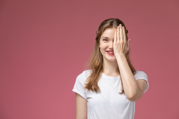 Vista frontal jovem mulher atraente em camiseta branca sorrindo e cobrindo metade do rosto na parede rosa modelo feminino jovem