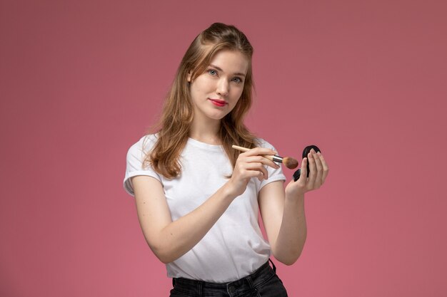 Vista frontal jovem mulher atraente em camiseta branca sorrindo e fazendo maquiagem na parede rosa modelo feminino pose foto colorida