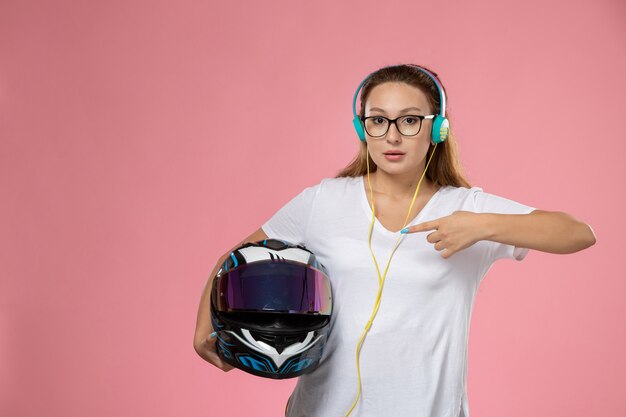 Vista frontal jovem mulher atraente em t-shirt branca casaco colorido ouvindo música segurando capacete no fundo rosa