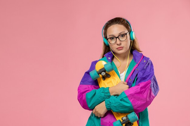 Vista frontal jovem mulher atraente em um casaco colorido de camiseta branca e ouvindo música na mesa rosa