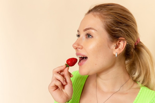 Vista frontal jovem mulher bonita com camisa verde comendo morango na parede creme claro fruta modelo mulher pose