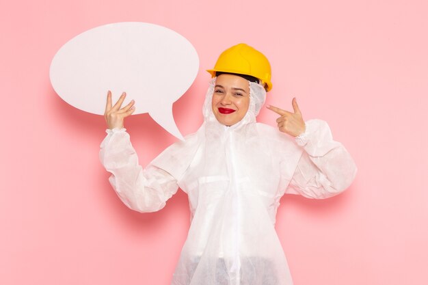 Vista frontal jovem mulher bonita em terno branco especial usando capacete protetor branco sinal com sorriso no espaço rosa mulher especial terno