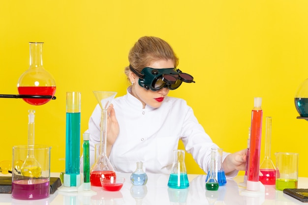 Vista frontal, jovem química feminina em um terno branco com soluções de ed, trabalhando com eles e sentada no espaço amarelo.