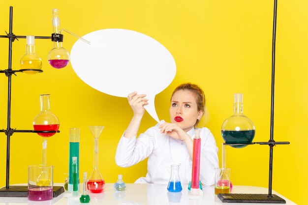 Vista frontal, jovem química feminina sentada em um terno branco com diferentes soluções, segurando uma placa branca sobre o processo de ciência química do espaço amarelo