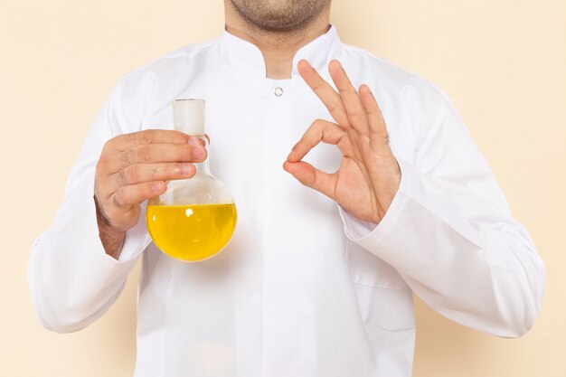 Vista frontal, jovem químico masculino, com terno especial branco, segurando o frasco com solução amarela na parede creme experimento de laboratório de ciências química científica