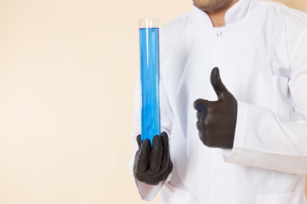 Vista frontal, jovem químico masculino em terno especial branco segurando um pequeno frasco com solução azul em experimento científico de química de laboratório de parede clara