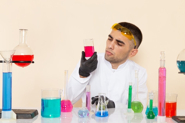 Vista frontal, jovem químico masculino em terno especial branco, sentado em frente à mesa com soluções, segurando o frasco e pensando na ciência da química do laboratório de mesa creme