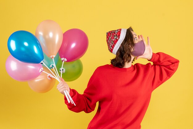 Vista frontal jovem segurando balões na máscara em amarelo celebração festa emoção ano novo cor mulher