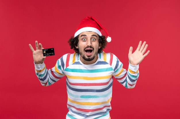 Vista frontal jovem segurando um cartão de banco preto na parede vermelha dinheiro vermelho masculino emoções férias