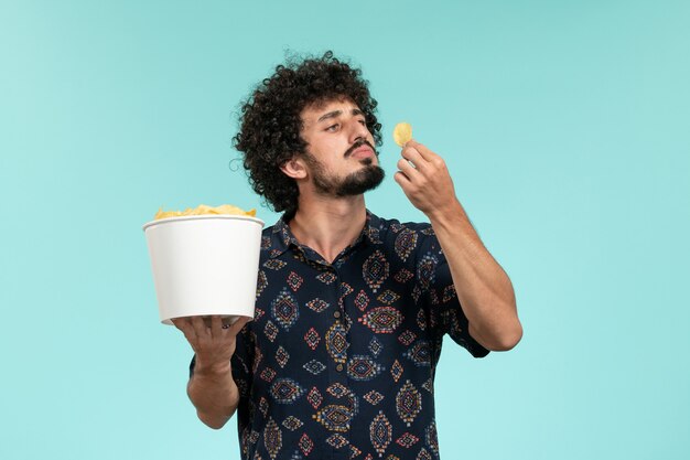 Vista frontal jovem segurando uma cesta com batatas cips e comendo na parede azul cinema filmes cinema teatro masculino