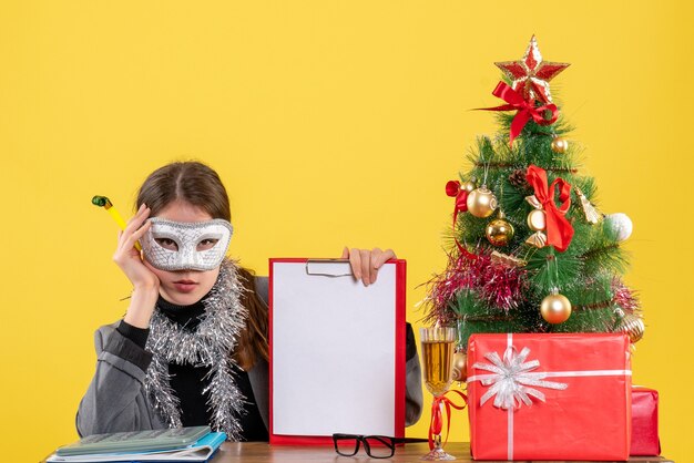 Foto grátis vista frontal jovem séria com máscara sentada à mesa segurando uma árvore de natal e coquetel de presentes