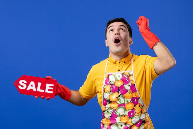 Vista frontal maravilhada governanta masculina com camiseta amarela segurando uma placa de venda, olhando para cima com surpresa no espaço azul