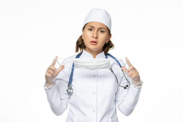 Vista frontal médica em traje médico branco segurando máscara devido a coronavírus no isolamento de vírus de doença pandêmica de parede branca