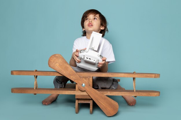 vista frontal menino bonito adorável segurando o controle remoto com avião de madeira em cima da mesa azul