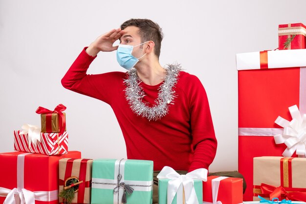 Vista frontal observando jovem com máscara sentado em volta de presentes de Natal