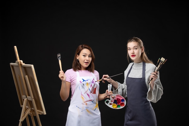 Vista frontal pintoras segurando tintas e borlas para desenhar na parede preta desenhar trabalho de pintura imagem arte foto artista a cores
