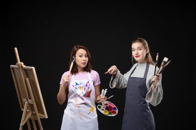 Foto grátis vista frontal pintoras segurando tintas e borlas para desenhar na parede preta desenhar trabalho de pintura imagem arte foto cores artista