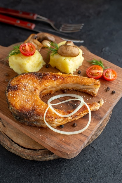 Vista frontal próxima de farinha de peixe frito com cogumelos, vegetais, queijo e talheres em uma placa de madeira na superfície preta angustiada