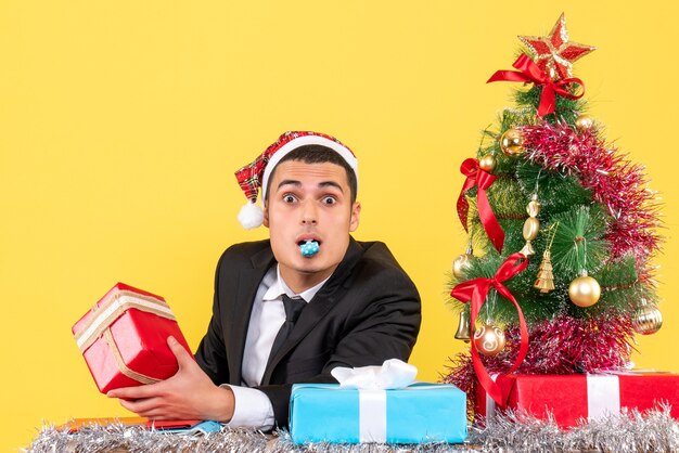 Vista frontal vagando homem com chapéu de Papai Noel sentado à mesa segurando presentes usando a árvore de natal do noisemaker e presentes