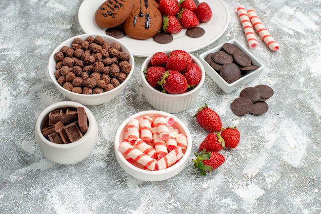 Vista inferior biscoitos morangos e chocolates redondos no prato oval branco tigelas com doces morangos chocolates cereais no fundo cinza-branco
