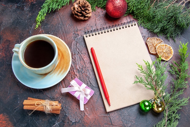 Vista inferior caneta vermelha um caderno pinheiro galhos de árvore de natal brinquedos bola de canela em pau uma xícara de chá em fundo vermelho escuro foto de natal