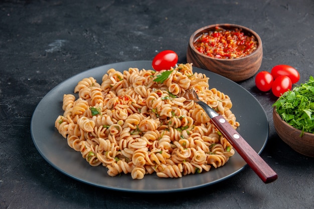 Vista inferior da massa rotini com garfo de tomate cereja no prato salsa e molho de tomate em tigelas tomate cereja na mesa escura