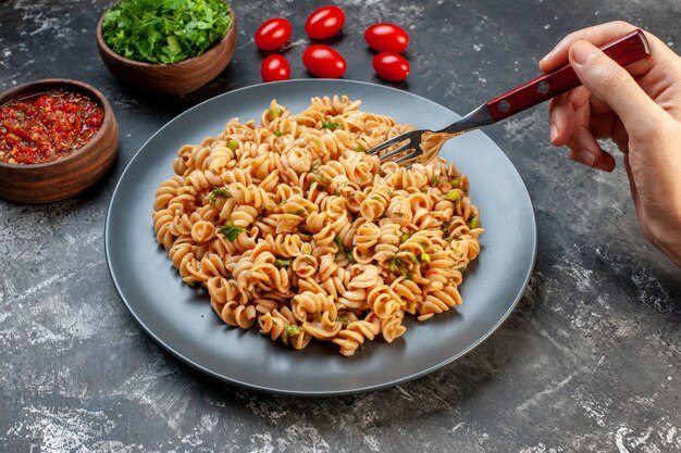 Vista inferior da massa rotini no garfo do prato em folhas verdes picadas à mão feminina e molho de tomate em tigelas na mesa cinza