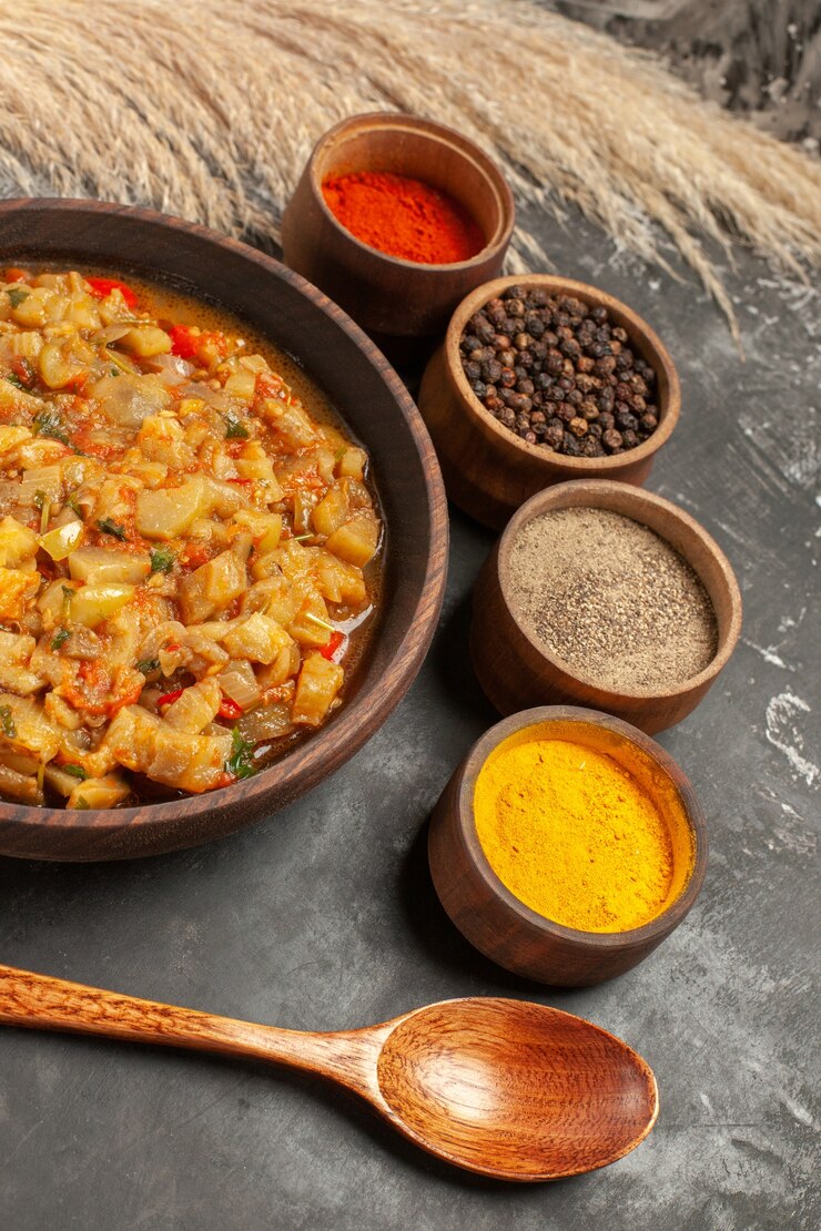 Foto grátis vista inferior da salada de berinjela assada em uma tigela diferentes especiarias em pequenas tigelas colher de pau na mesa escura