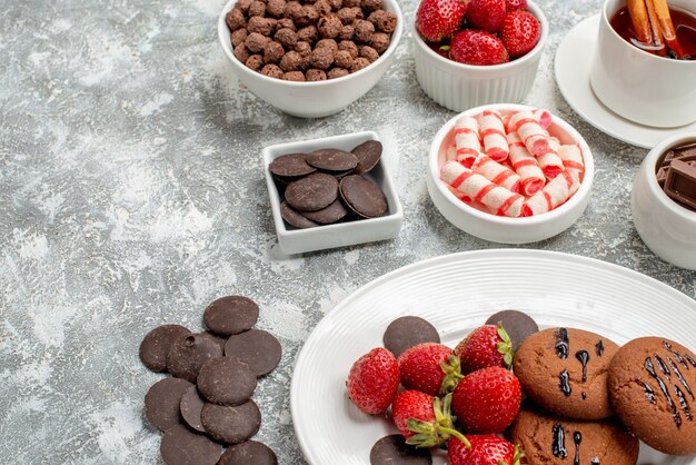 Vista inferior de perto cookies, morangos e chocolates redondos no prato oval taças com doces, morangos, chocolates, cereais e chá de canela na mesa branco-acinzentada