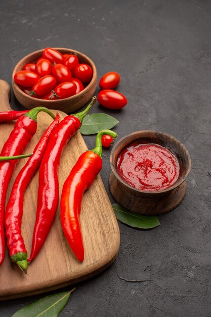 Vista inferior de perto uma tigela de tomate cereja, pimentão vermelho quente na tábua de cortar folhas de louro e uma tigela de ketchup na mesa preta