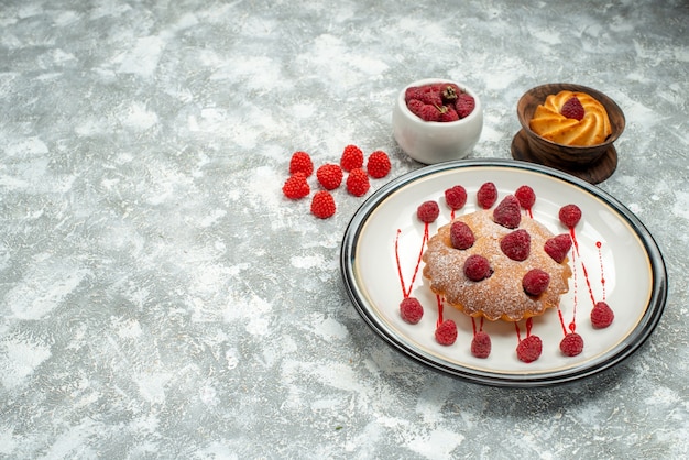 Vista inferior do bolo de frutas vermelhas em um biscoito de prato oval branco em uma tigela de madeira framboesas em uma tigela em uma superfície cinza espaço livre
