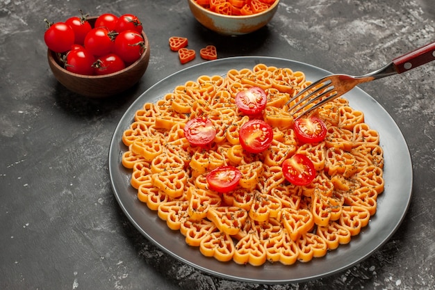 Vista inferior dos corações de massa italiana cortam tomate cereja no prato, garfo, tomate cereja e macarrão coração em uma tigela na mesa cinza