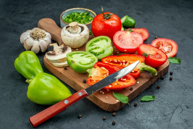 Vista inferior legumes pepinos tomates verdes e vermelhos faca pimentão na tábua de cortar alho verdes em uma tigela na mesa preta