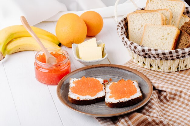 Vista lateral caviar vermelho torrada pão de centeio com queijo cottage vermelho caviar manteiga pão branco laranja e banana na mesa branca
