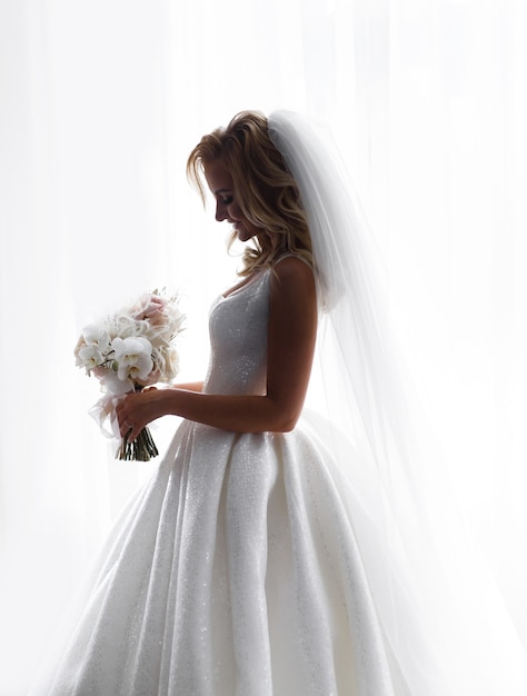 Vista lateral da adorável noiva, usando um vestido de noiva brilhante e véu, considerando as flores