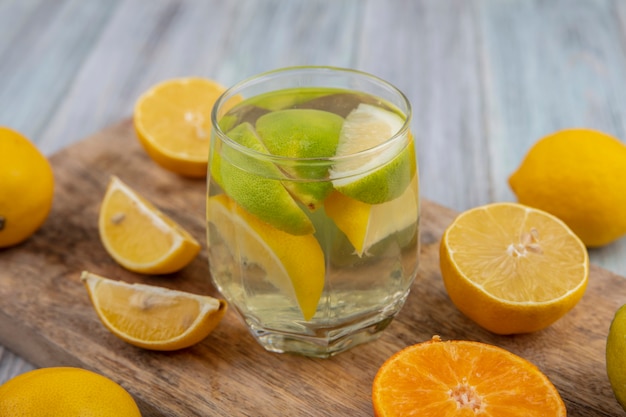 Vista lateral da água desintoxicante em um copo com fatias de limão e meia laranja e limão em uma tábua de cortar
