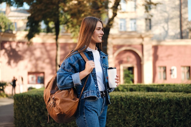 Vista lateral da estudante casual na jaqueta jeans com mochila e café olhando alegremente para longe no campus universitário ao ar livre