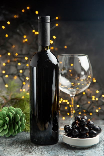 Vista lateral da garrafa de vinho tinto para a celebração de um copo vazio e uma casquinha de uva preta em fundo escuro