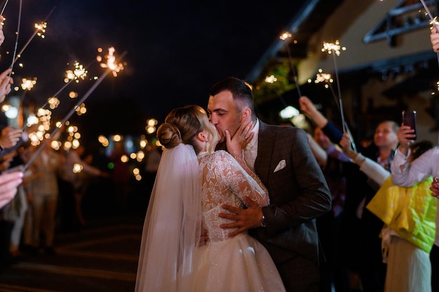 Vista lateral da linda noiva e do noivo que se abraçam e se beijam enquanto os convidados seguram brilhos frios que queimam e criam arco brilhante durante a cerimônia noturna ao ar livre