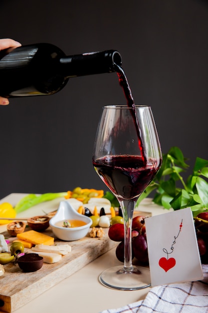 vista lateral da mão de uma mulher derramando vinho tinto no copo e queijo azeitona noz uva e amor cartão na superfície branca e parede preta