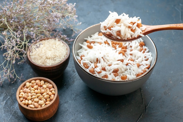 Vista lateral da mão tomando uma saborosa refeição de arroz com uma colher de pau com feijão em uma panela pequena branca e seus ingredientes sobre fundo azul